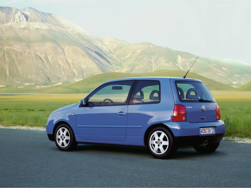 Volkswagen Lupo azul visto de costado en una carretera frente a las montañas