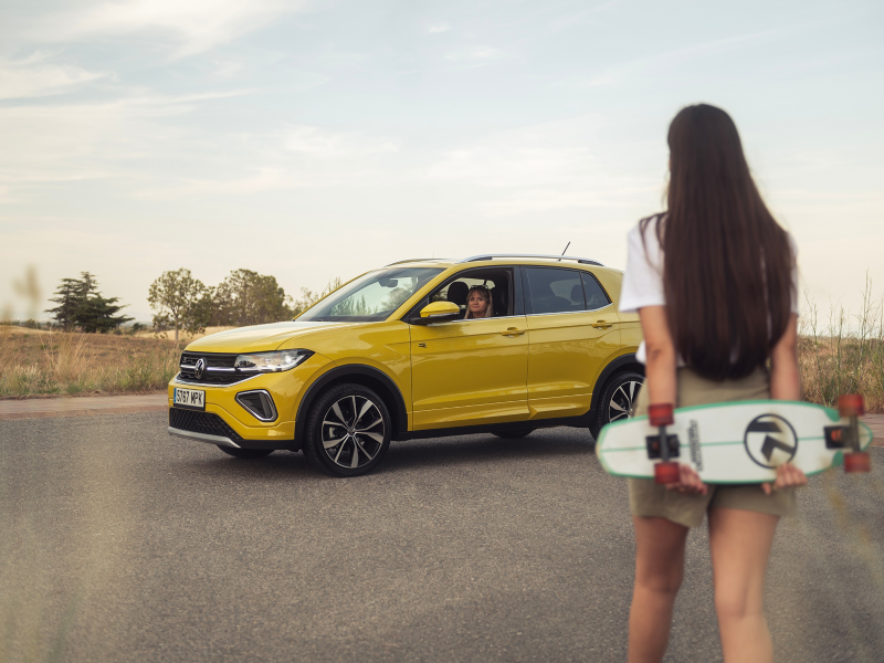 Una mujer en el interior de un Volkswagen T-Cross de color amarillo delante de una niña con un skateboard