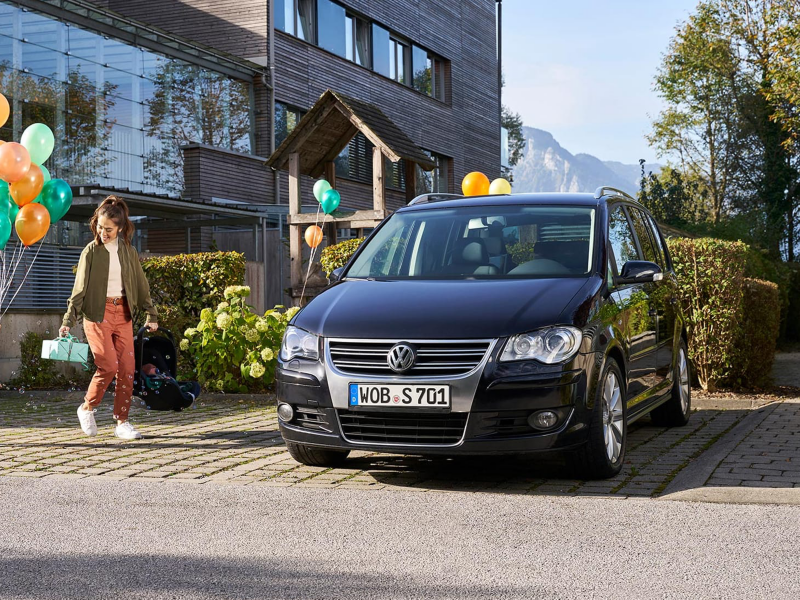 Una chica con globos y un regalo acercándose a un Volkswagen Touran negro