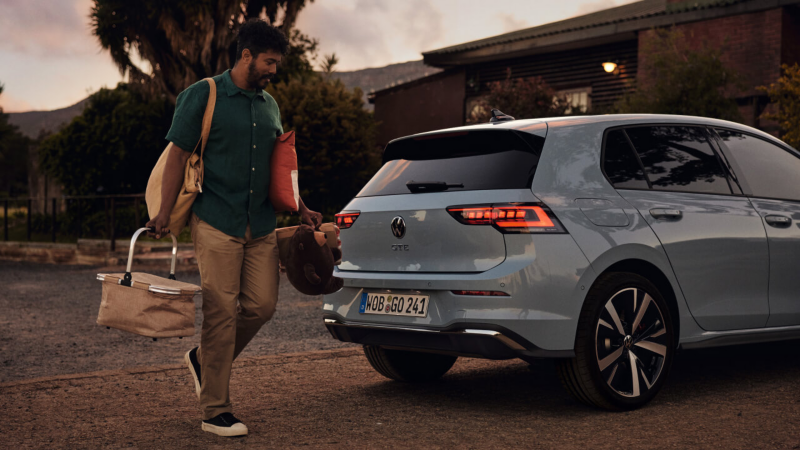Un chico cargado con bolsas detrás de un Volkswagen Golf GTE estacionado frente a una casa al atardecer 