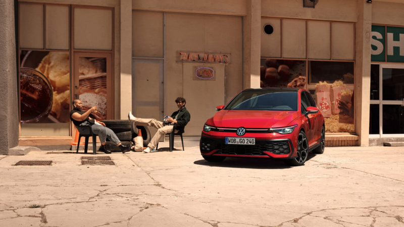Un Volkswagen GTI de color rojo estacionado fuera de un establecimiento y dos personas sentadas