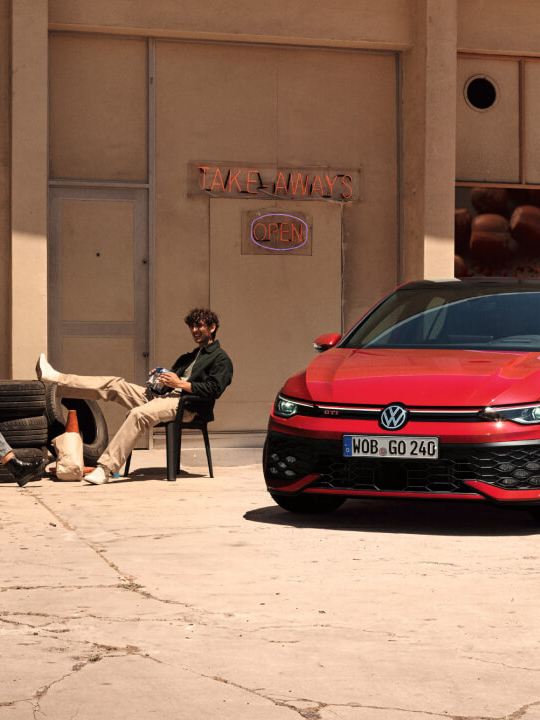 Dos personas sentadas en una silla junto a un Golf GTI de color rojo estacionado fuera de un estableicmiento