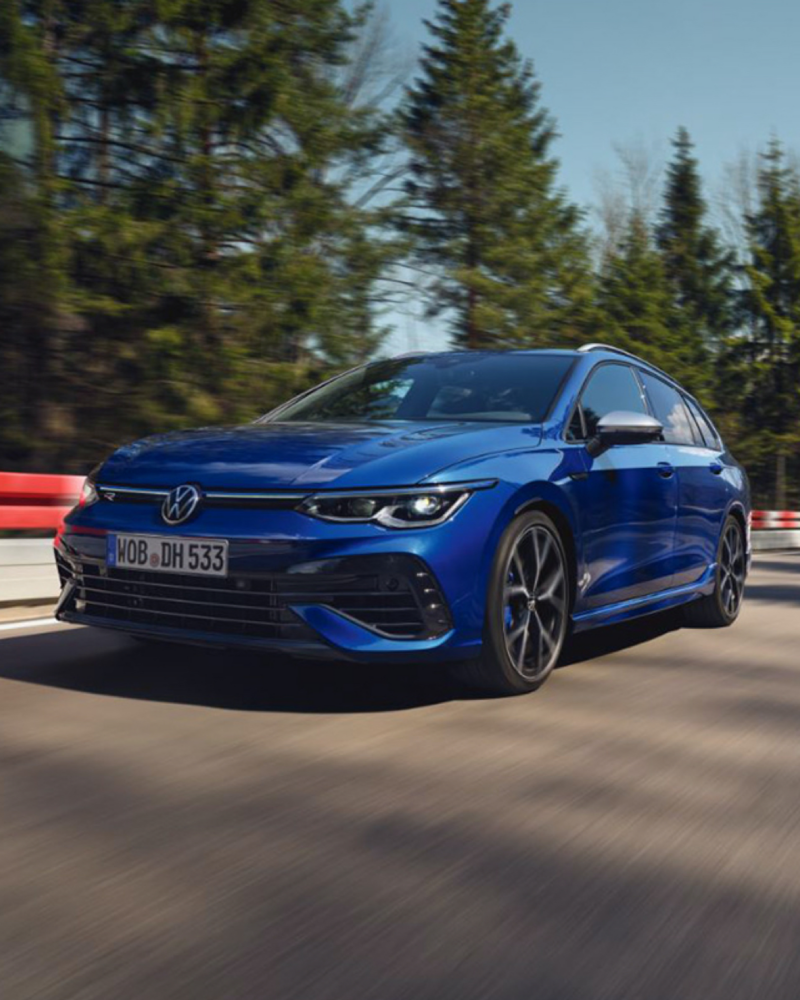 Vista frontal de un Golf 8 Variant R azul en una autopista con pinos de fondo