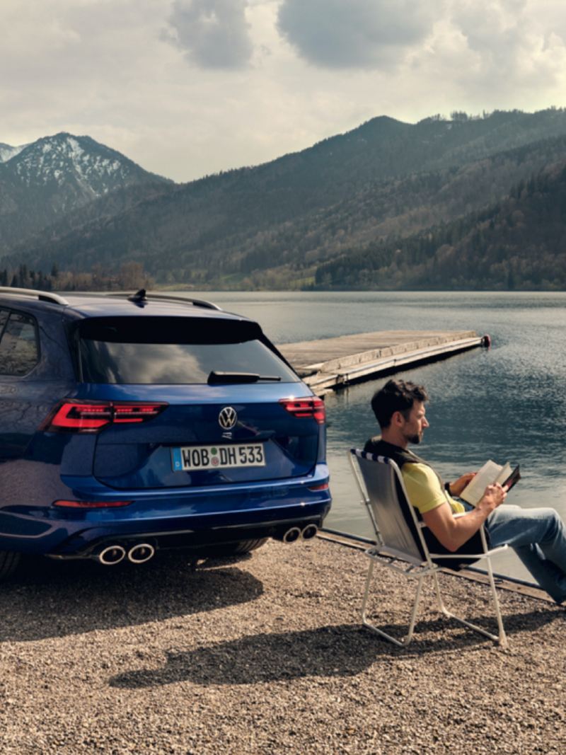 Un hombre leyendo sentado en una silla plegable frente a un lago junto a un Volkswagen Golf 8 Variant R azul
