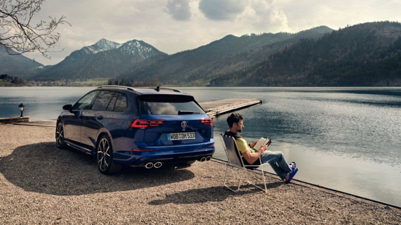 Un hombre leyendo sentado en una silla plegable frente a un lago junto a un Volkswagen Golf 8 Variant R azul