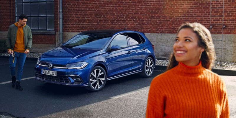 Un Volkswagen Polo azul aparcado mientras un chico camina por delante y una chica en primer plano