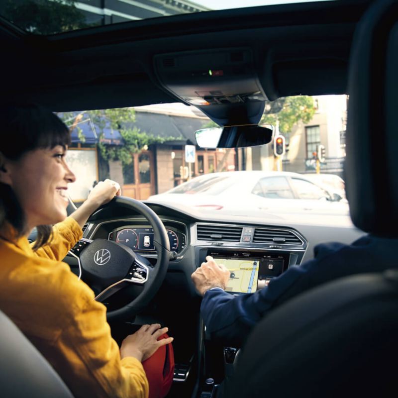 Dos personas dentro de Volkswagen Tiguan azul enfocando de atrás hacia adelante. La persona de la izquierda está cogiendo el volante y la de la derecha tocando la pantalla con el dedo.