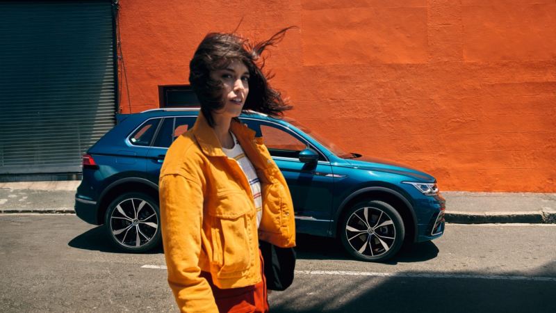 Chica con chaqueta amarilla delante de un Volkswagen Tiguan azul aparcado frente a un muro naranja