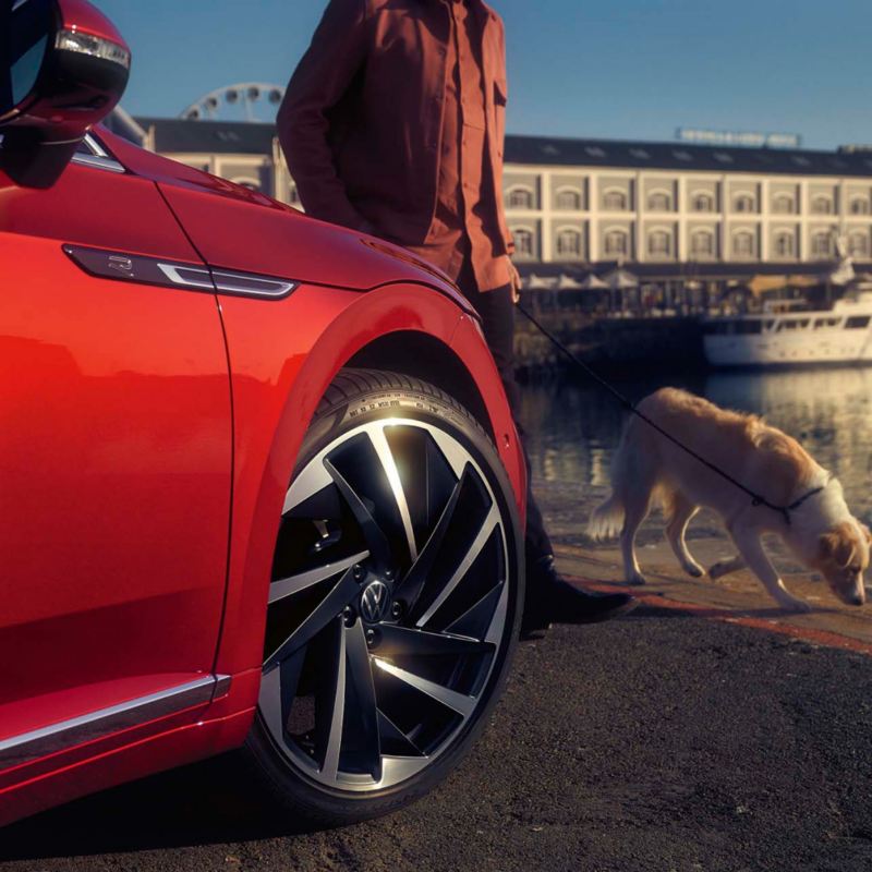 Hombre paseando con un perro delante de un Volkswagen Arteon Shooting Brake rojo