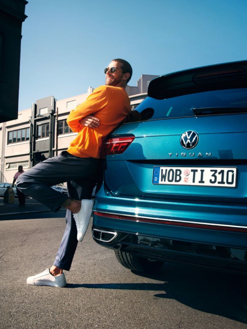 Hombre joven sonriendo apoyado en la parte trasera de un Volkswagen Tiguan azul metalizado aparcado