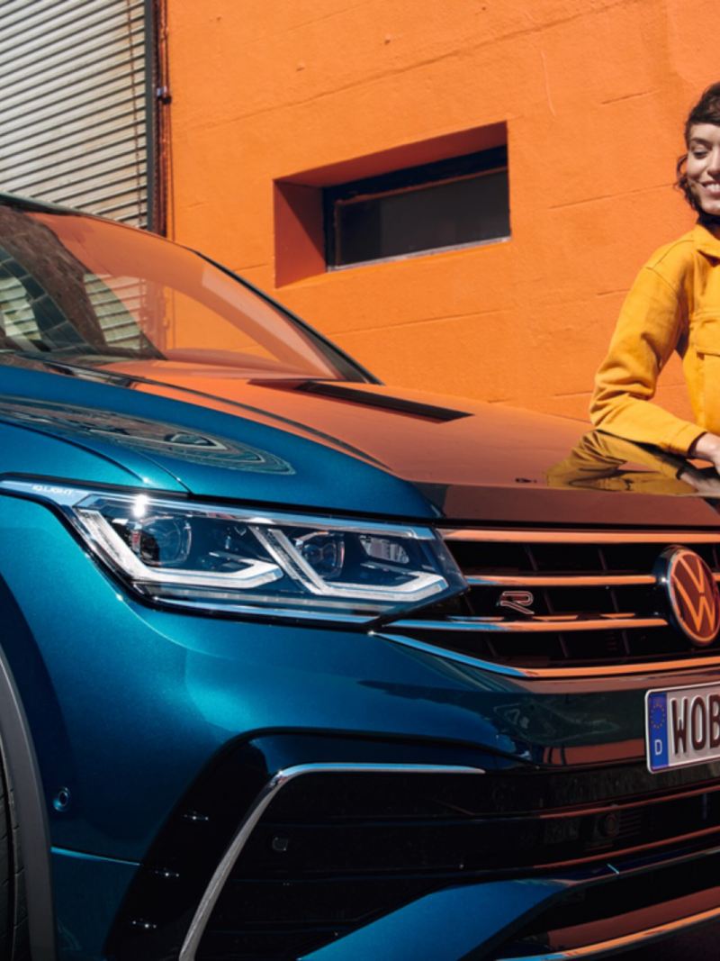 Chica sonriendo apoyada sobre el capó de un Volkswagen Tiguan azul metalizado a aparcado en la calle