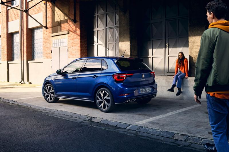 Una chica junto a un Volkswagen Polo de color azul estacionado en la calle