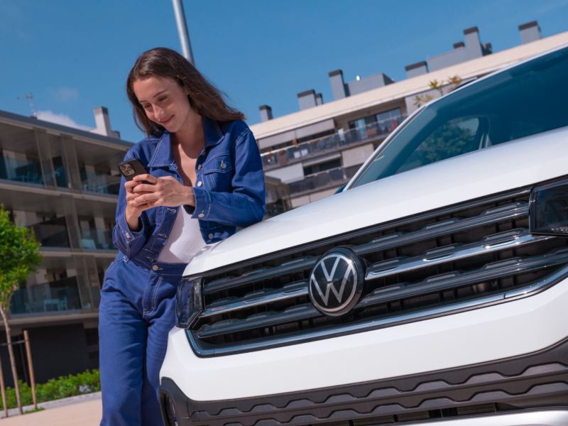 Chica con un móvil apoyada en el capó de un Volkswagen T-Cross blanco bajo el sol