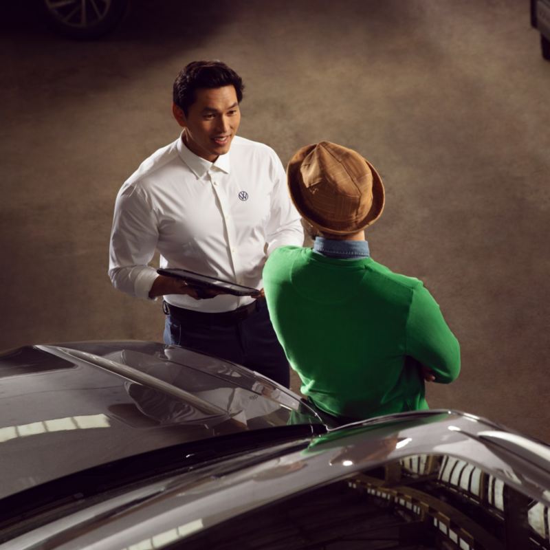 Mecánico hablando con un hombre joven apoyado en el capó de un Volkswagen visto desde arriba