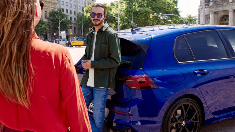 Chico apoyado en la parte posterior de un Golf R azul, mientras mira a una chica