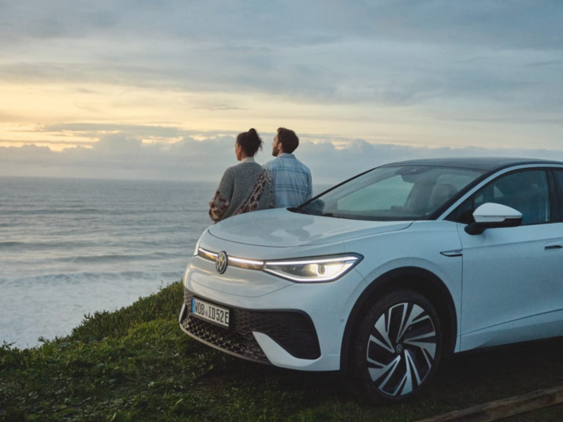 Una pareja apoyada en un Volkswagen ID.4 blanco frente al mar al atardecer