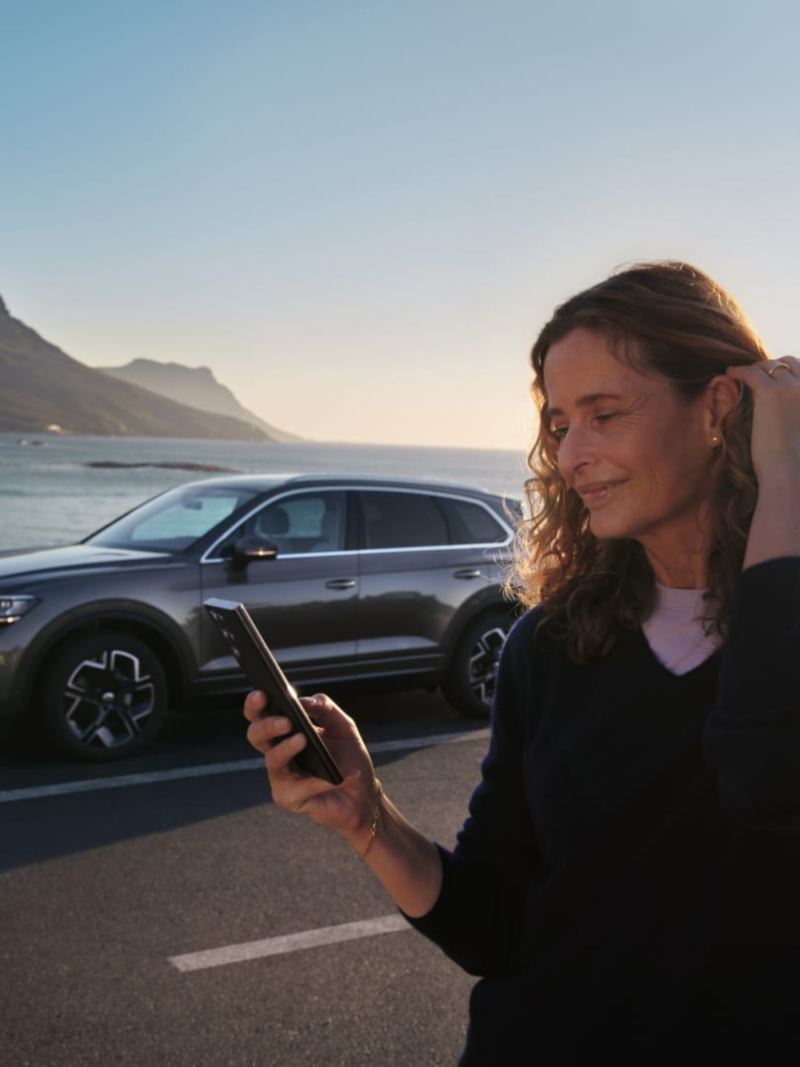 Una mujer con un móvil de pie delante de un Volkswagen junto al mar