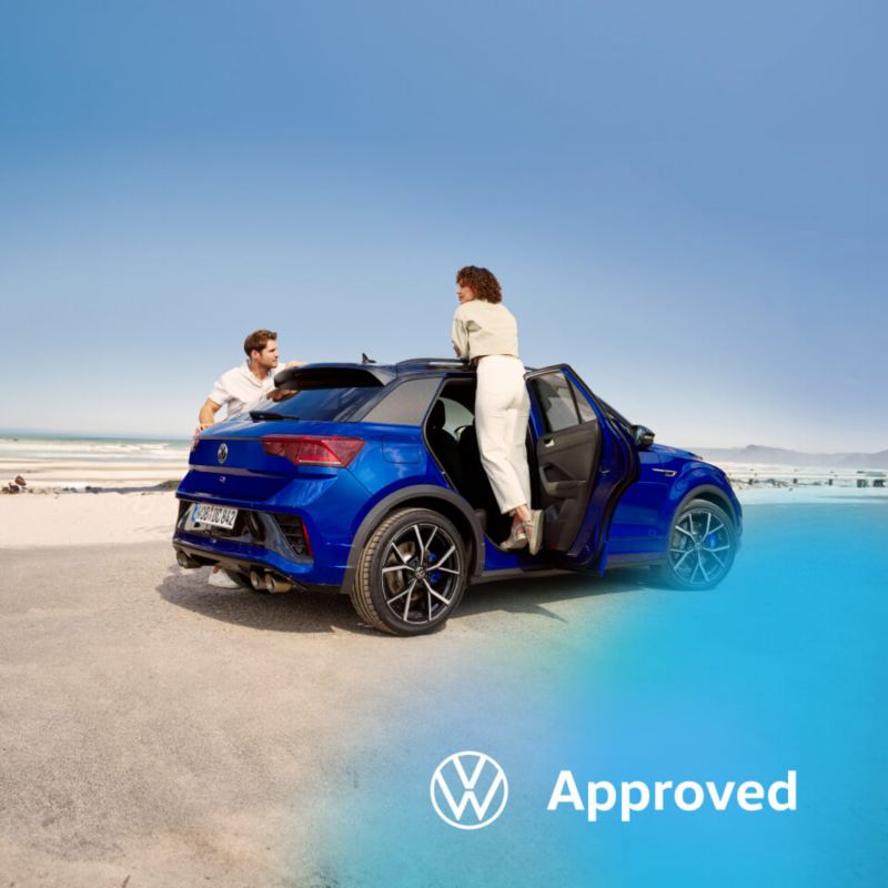 Una pareja en un Volkswgen de color azul oscuro estacionado en la playa con el logo Approved