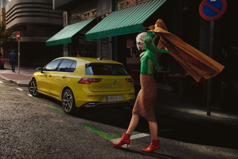 Una mujer elegante camina hacia un Volkswagen Golf amarillo 