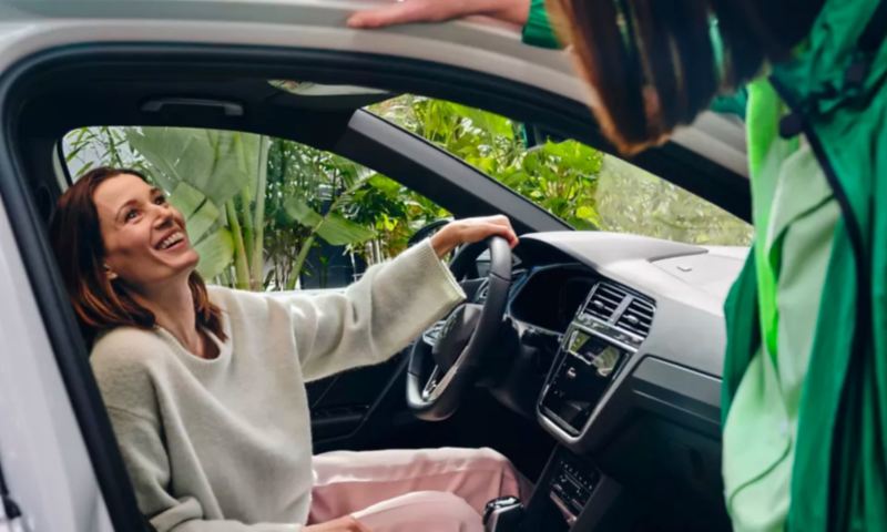 Chica sonriendo sentada dentro de un Volkswagen Tiguan híbrido enchufable hablando con otra que está fuera