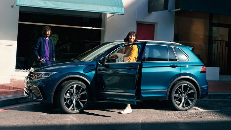 Una chica junto a un Volkswagen Tiguan azul visto de costado aparcado en una calle