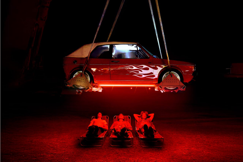 Tres personas tumbadas con iluminación roja bajo un Golf MK1 descapotable rojo con llamas blancas