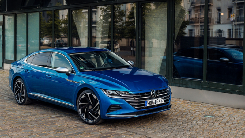 Front view of a bright blue Volkswagen Arteon parked on a cobblestone street in front of a building with large glass windows.