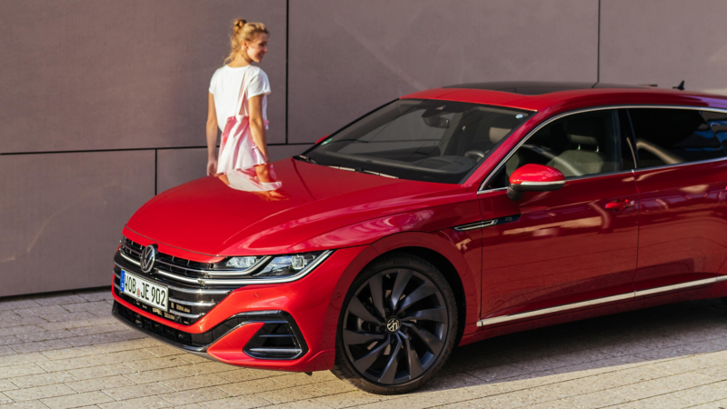 A front three-quarter view of a red Volkswagen Arteon Shooting Brake parked on a paved area, with a woman walking in the background.