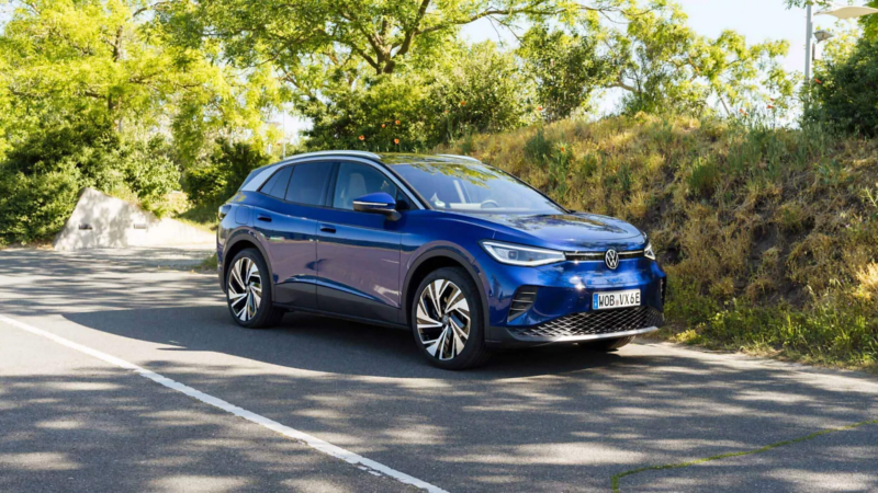 A front view of a metallic blue Volkswagen ID.4 parked on an asphalt road surrounded by green trees and a grassy hillside.