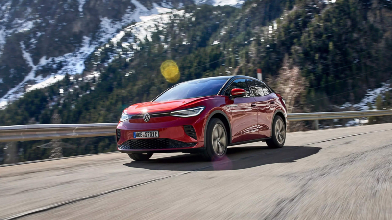 A red Volkswagen ID.5 driving on a mountain road with snow-dusted slopes and green pine trees in the background.