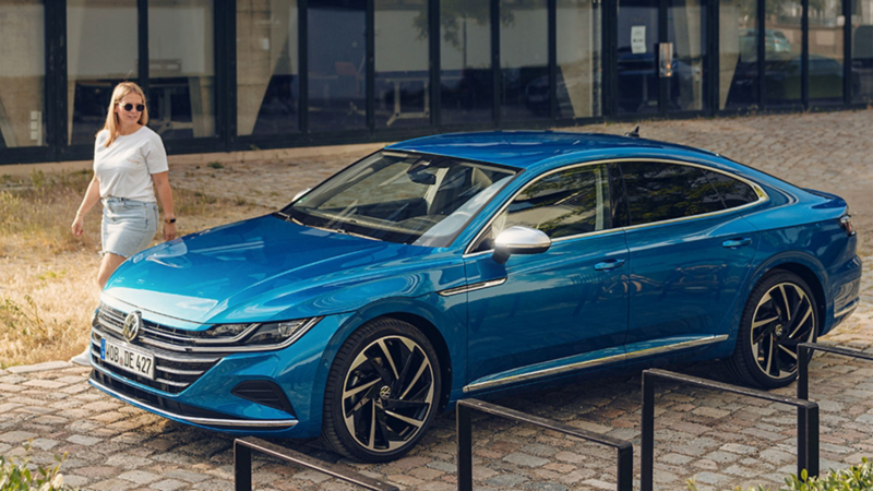 A blue Volkswagen Arteon sedan parked on a cobblestone area, with a woman walking in the background near a glass building.