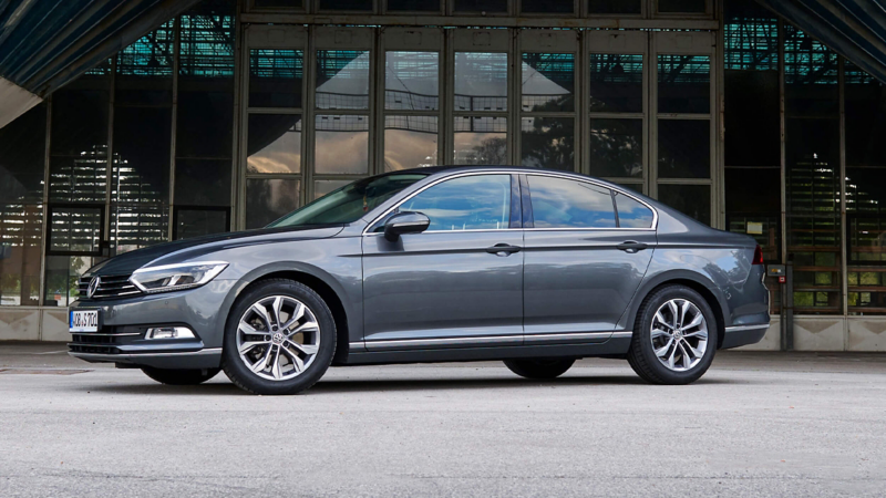 A side profile of a dark grey Volkswagen Passat parked in front of a building with glass doors and a metal structure.