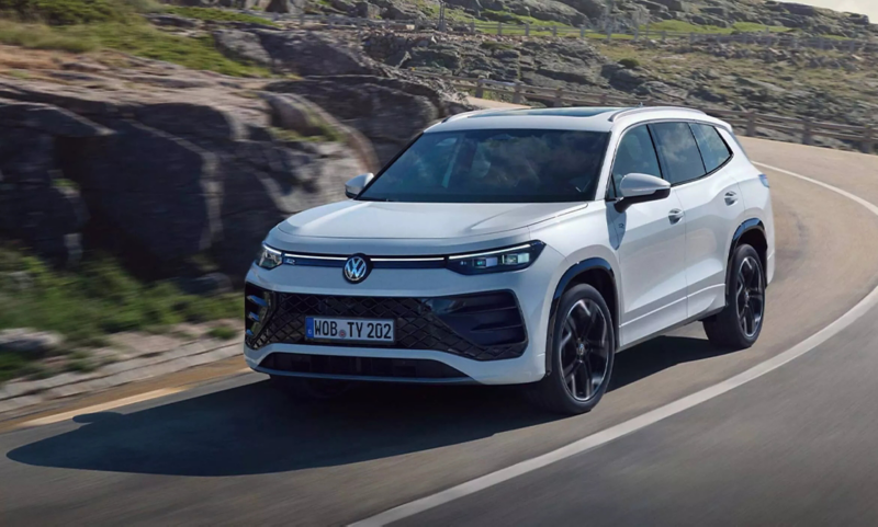 A white Volkswagen Tayron SUV driving on a winding coastal road with rocky hills in the background.