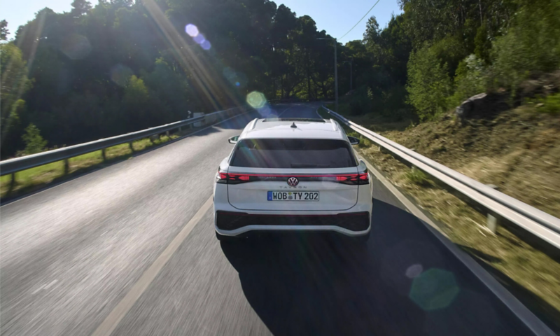 Rear view of a white Volkswagen Tayron driving on a sunny road lined with trees.