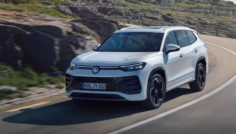 A white Volkswagen Tayron SUV driving on a winding coastal road next to a rocky hillside.