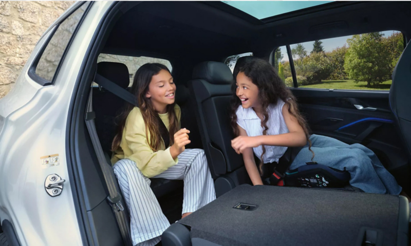 Two girls smiling and talking in the rear seats of a vehicle with a panoramic sunroof and folded row.
