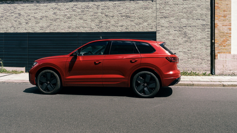 A red Volkswagen Touareg SUV parked on a city street, viewed from the side profile against a brick building background.