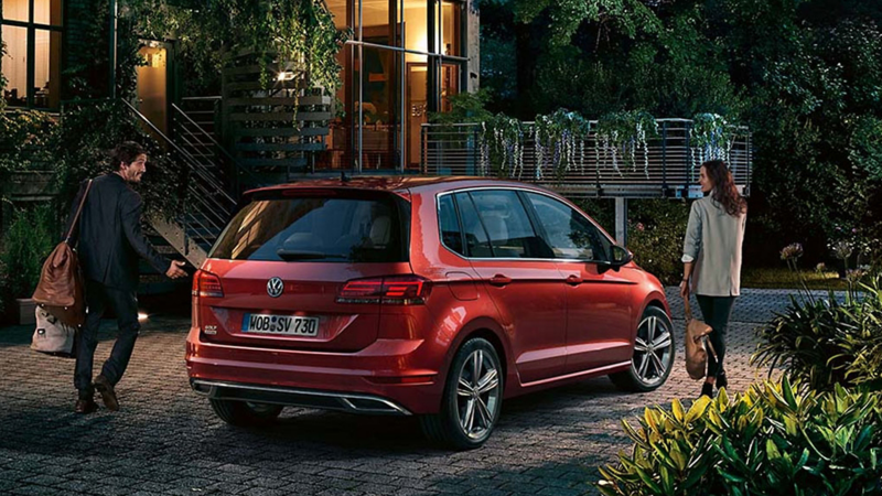 Rear shot of a red Volkswagen Golf SV, outside a hotel a couple leave with their luggage. 