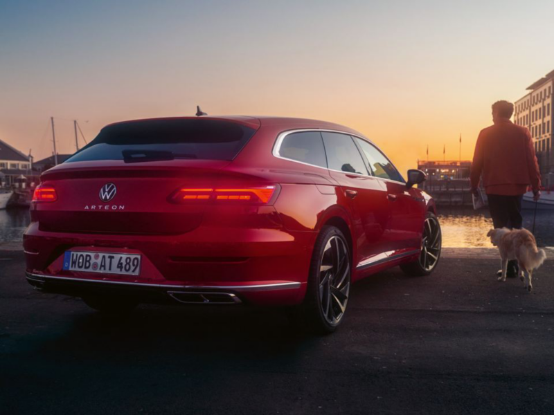 A tail light view of a red Arteon Shooting Brake parked at a dock, with a man and dog walking alongside it.