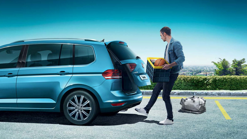 A man with a box using the hands-free sensors to open the boot of a blue Volkswagen Touran.