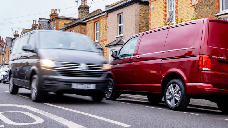 A VW van driving down a street next to parked vehicles in a suburban setting.