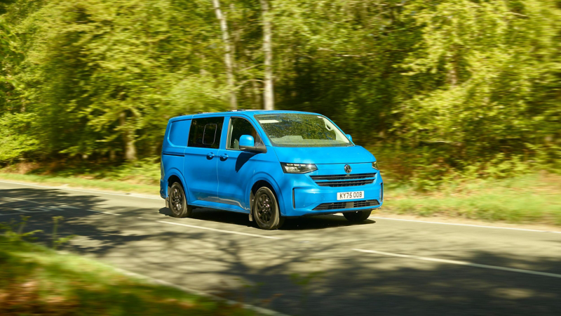 A blue van drives down a sunlit, tree-lined road, surrounded by lush green foliage. The scene conveys a sense of motion and tranquility.