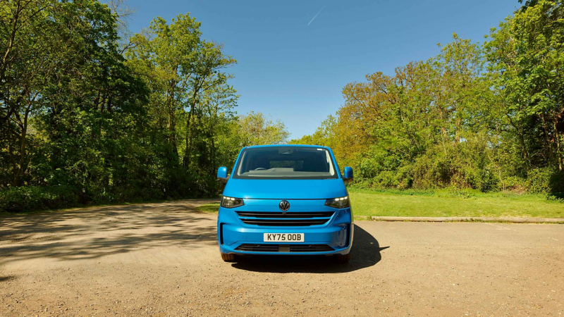 A bright blue van is parked on a sunlit, empty dirt road surrounded by lush green trees under a clear blue sky. The scene feels calm and serene.