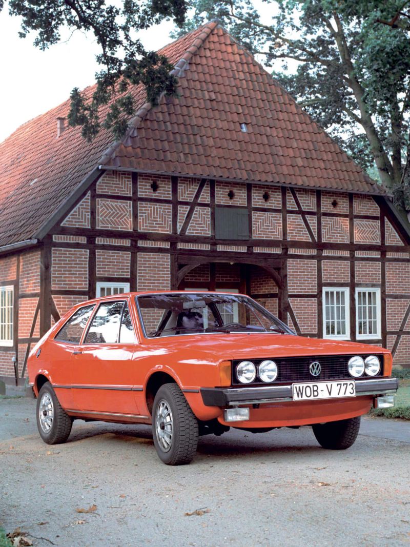 Un VW Scirocco rouge, première génération, devant une maison traditionnelle allemande.