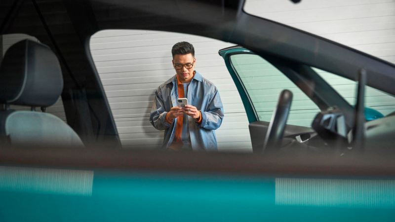 Un homme utilise son smartphone. Nous le voyons au travers d'une vitre de Volkswagen au tout premier plan.