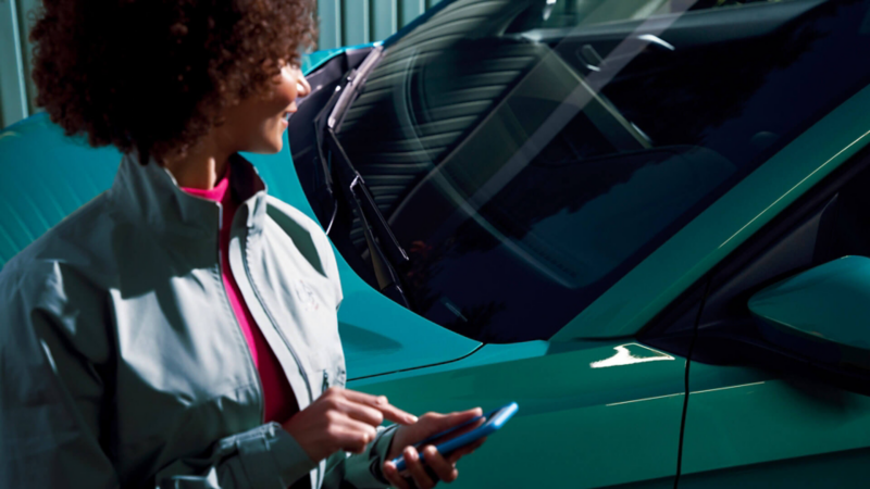 Gros plan sur une femme qui se tourne pour regarder son VW T-Cross, alors qu'elle manipule son smartphone.