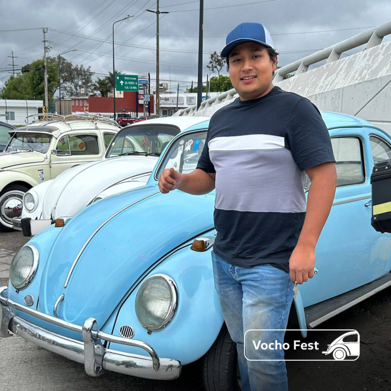 Conductor de Vocho posa delante de un Volkswagen sedán de color azul. 