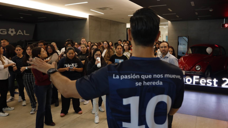 	Hombre da discurso a multitud junto a Volkswagen Sedan de color rojo en concesionaria.