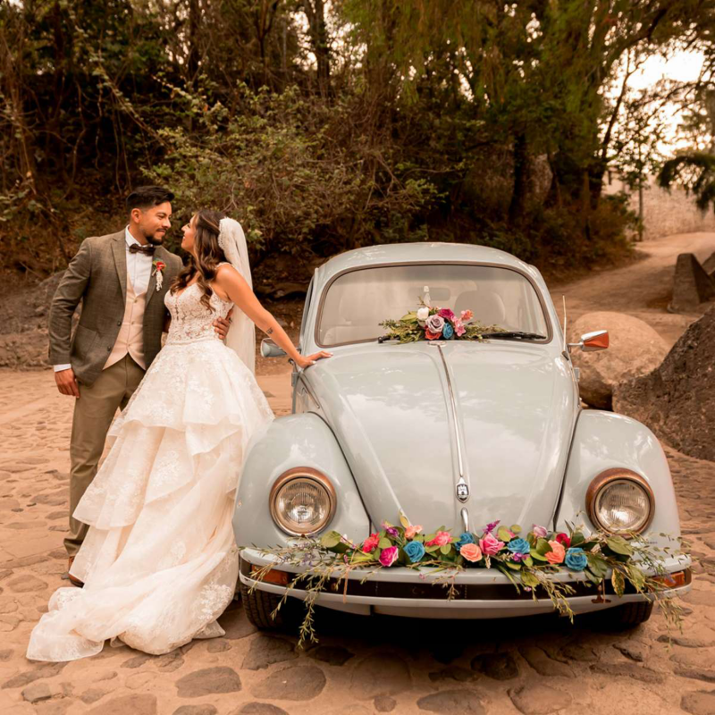 Pareja de novios posa junto a Volkswagen Sedán clásico de color azul claro con arreglo floral.
