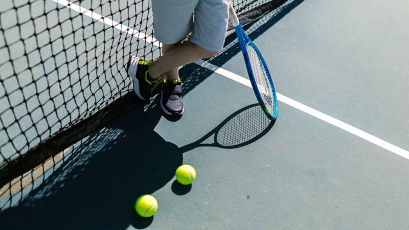 Hombre parado a lado de una red de tenis en cancha, con una raqueta y pelotas.